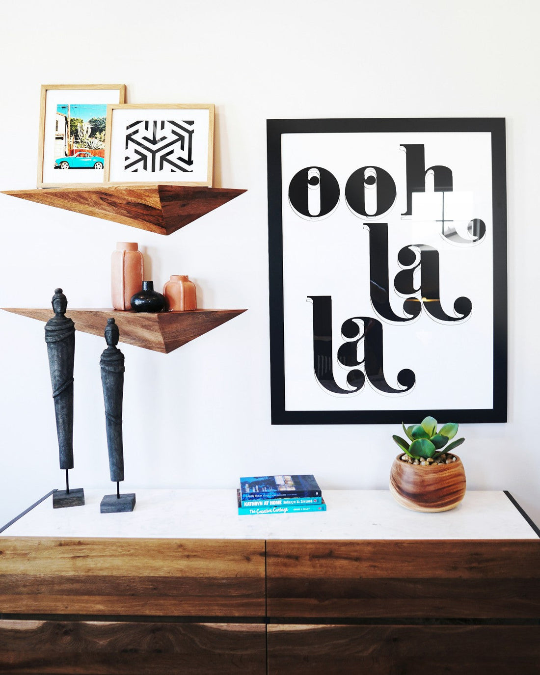 A selection of home accessories sitting on a chest of drawers and angular shelves, as well as a print that says 'ooh la la'