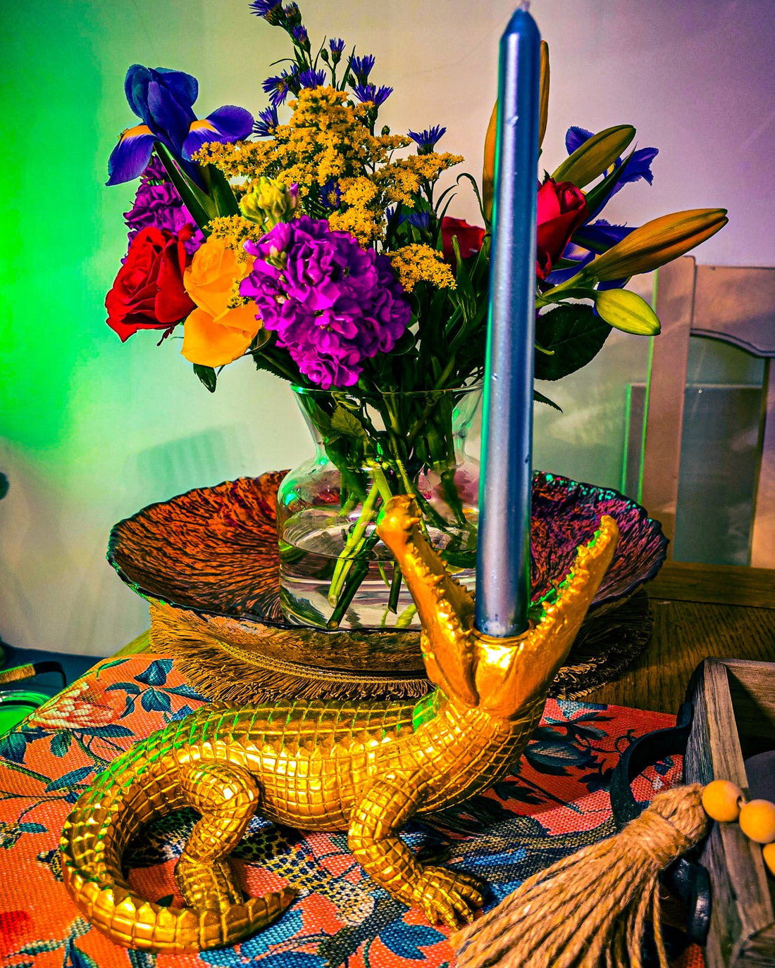 a gold crocodile candle holder with a blue dinner candle in its mouth, with bright coloured fresh flowers in a vase in the background